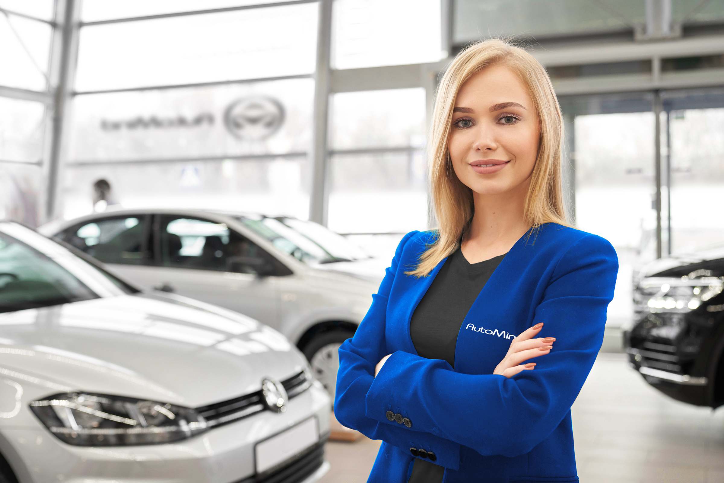 2A4TEFG Front view of attractive female car dealer with folded arms standing in auto salon, looking at camera and posing. Clever worker selling new automobiles and helping customers in showroom.